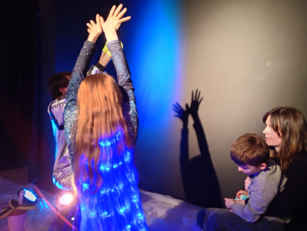 Aktorzy unoszą do góry ręce, tworząc na ścianie teatr cieni, obok siedzi chłopiec z opiekunką.