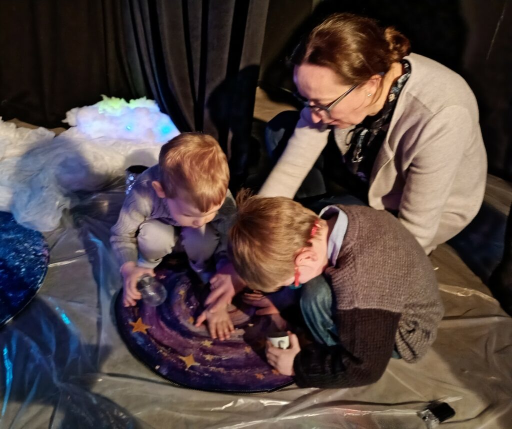 Dwaj chłopcy dotykają elementu scenografii umieszczonego na podłodze, obok wychowawczyni.