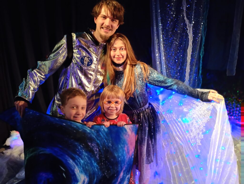 Chłopiec i dziewczynka trzymają przed sobą element dekoracji, za nimi stoją aktorzy 