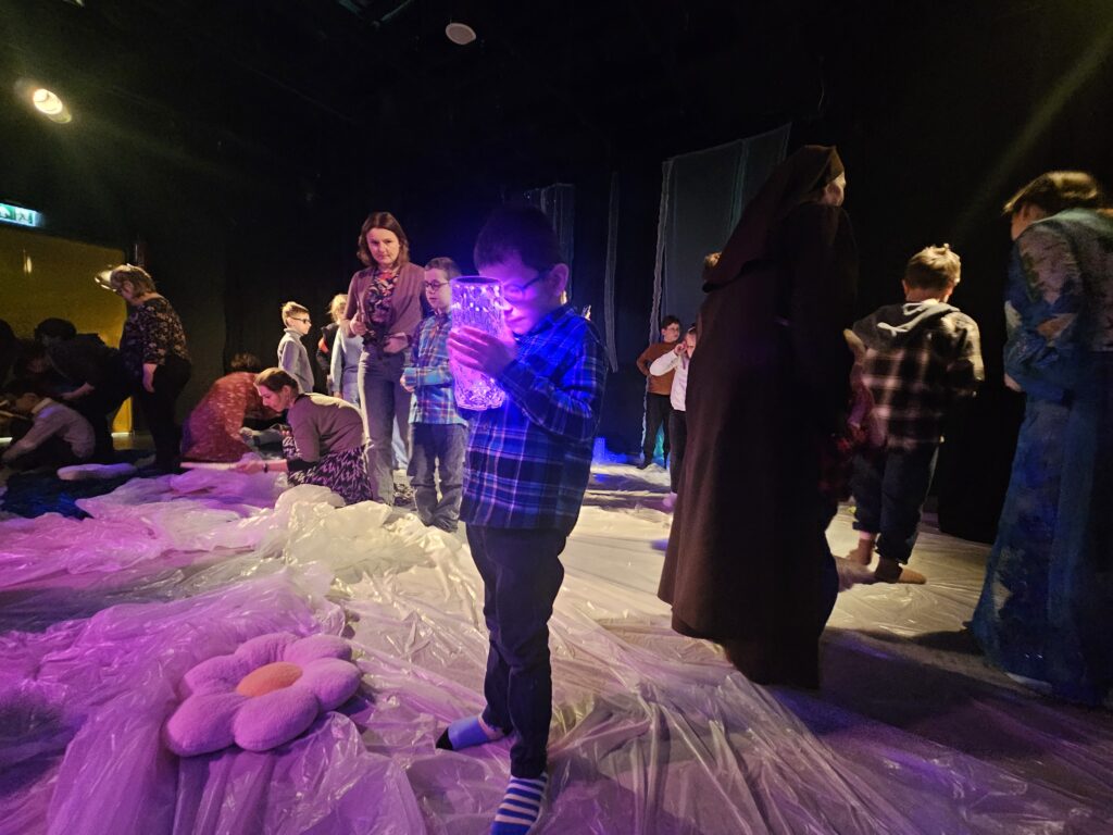 Chłopiec stojąc na scenie trzyma w rękach święcącą lampę, za nim dzieci wraz z opiekunami poznają scenografię.