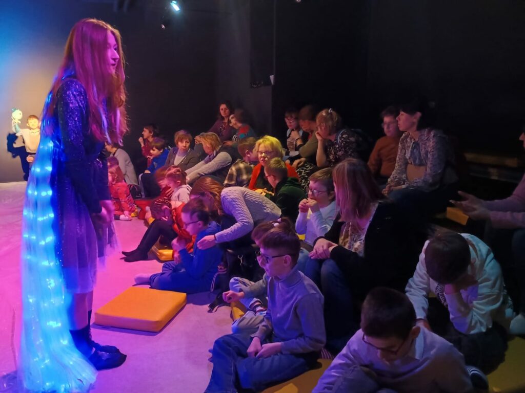 Dzieci i wychowawcy siedzą na podłodze, przed nimi stoi aktorka w  świecącej pelerynie.