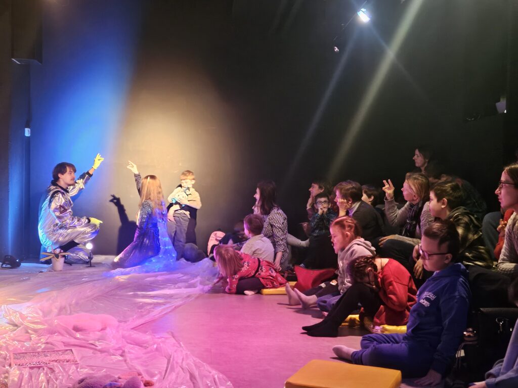 Dzieci i opiekunowie siedzą na podłodze, przed nimi aktorzy z uniesionymi rękoma, tworzą na ścianie cienie.