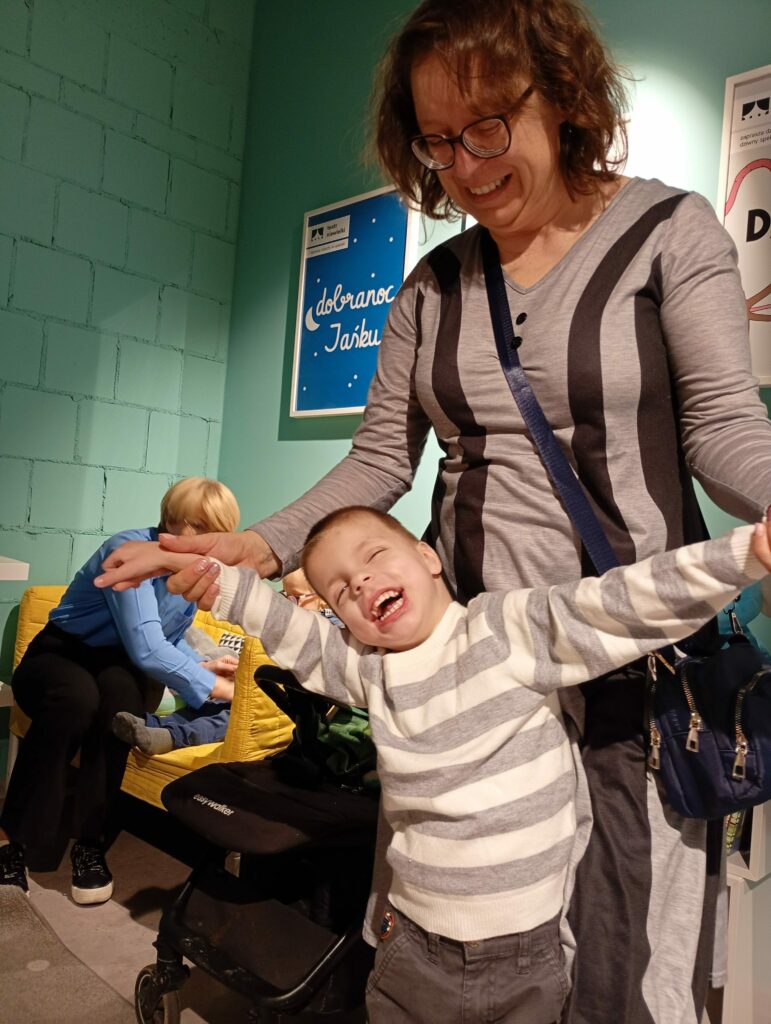 Uśmiechnięty chłopiec wyciąga ręce w bok, za nim opiekunka.