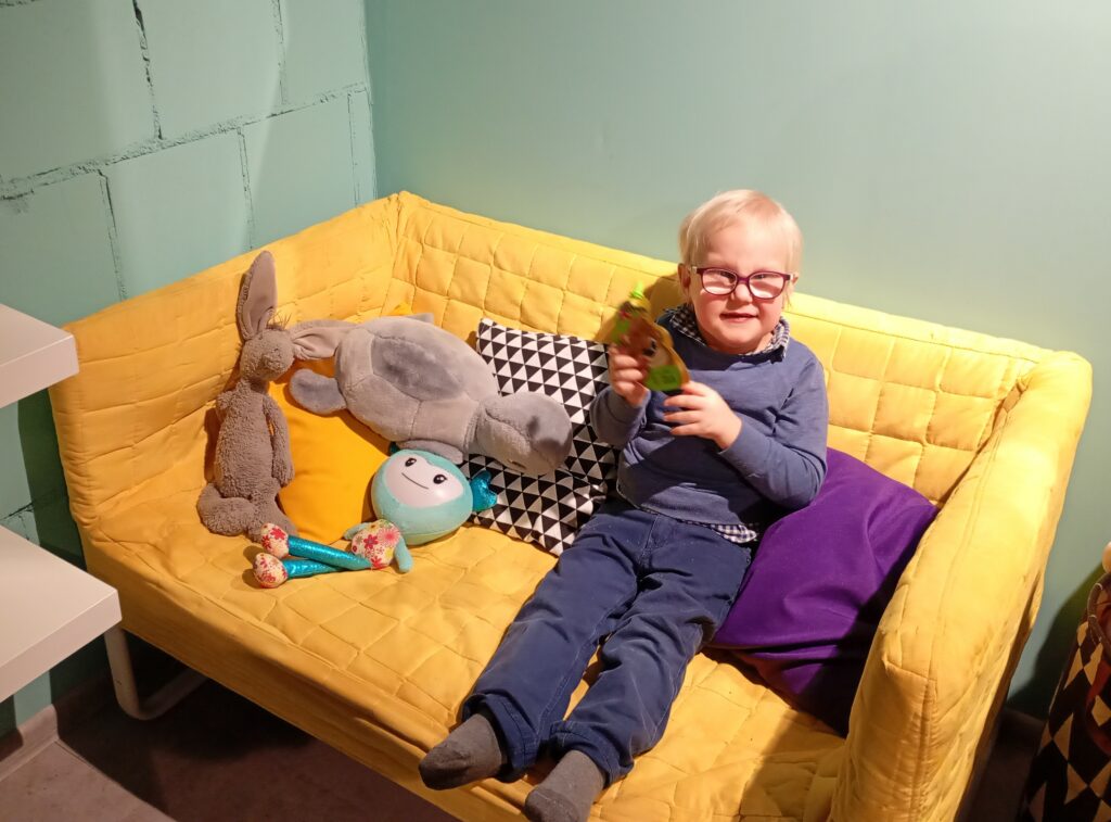 Chłopiec siedzi na kanapie, w rękach trzyma opakowanie po musie owocowym.