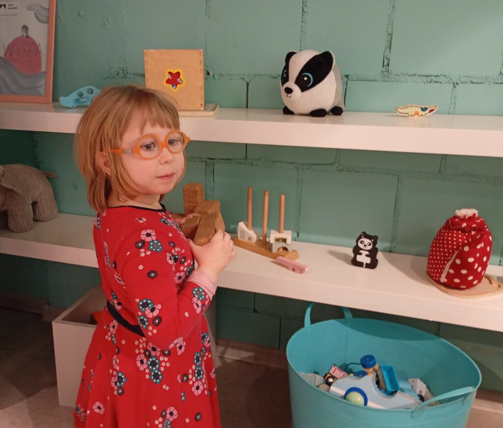 Dziewczynka stoi obok półek z zabawkami, w rękach trzyma klocki.