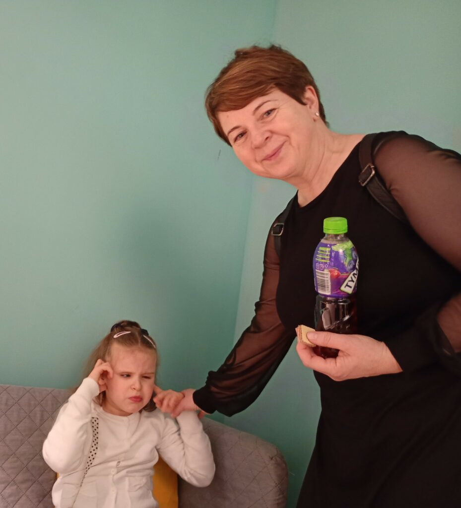 Dziewczynka siedzi na kanapie, obok stoi opiekunka. 