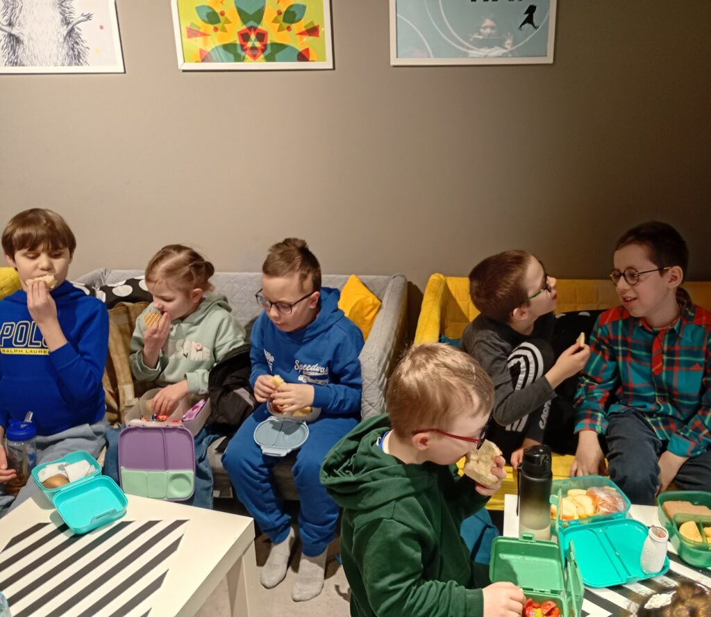 Chłopcy i dziewczynka siedzą na kanapach jedząc i rozmawiając. 