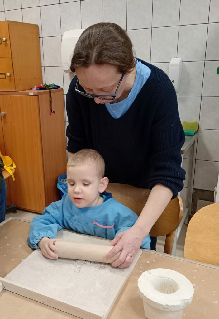 Uśmiechnięty chłopiec w fartuszku siedzi przy stole w pracowni ceramicznej. Przed nim na stole leży drewniana podkładka a na niej drobniutkie kawałki suchej gliny. Chłopiec z pomocą nauczycielki rozgniata kawałki gliny za pomocą wałka. 