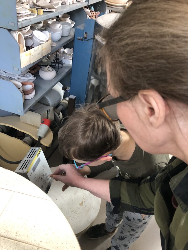 Chłopiec pochyla się nad programatorem pieca. Obok stoi nauczycielka i palcem chłopca naciska przycisk na programatorze. W tle widać regały z pracami z gliny.