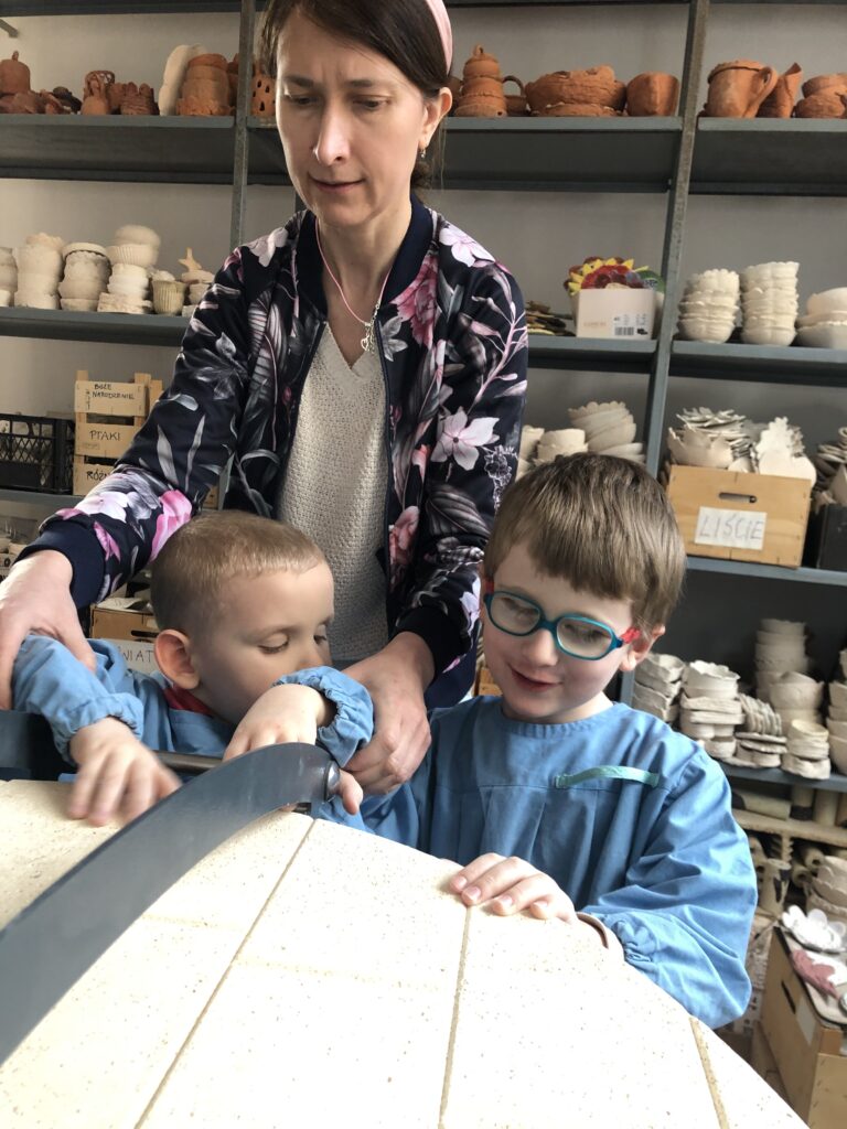 Dwóch chłopców stoi z nauczycielką przy wielkim piecu w kształcie walca.  Dotykają dłońmi pokrywy pieca. 