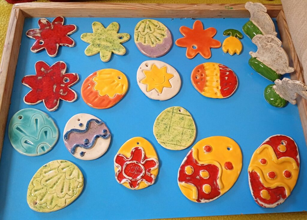 Na niebieskiej drewnianej podkładce leżą kolorowe ceramiczne prace: płaskie kwiatki i pisanki oraz stojące zajączki.