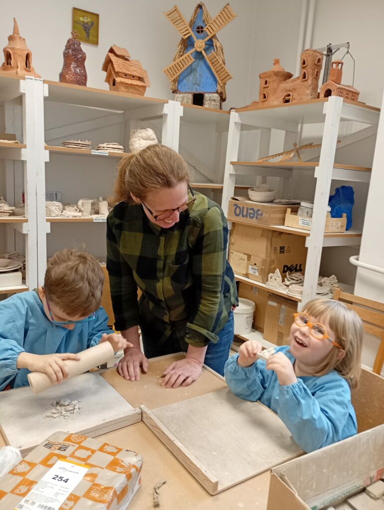 Chłopiec i dziewczynka w fartuszkach siedzą przy stole w pracowni ceramicznej. Przed każdym na stole leży drewniana podkładka. Na podkładce chłopca leżą drobne kawałki suchej gliny. Chłopiec trzyma w dłoniach wałek uniesiony nad gliną. Dziewczynka trzyma w dłoniach kawałek gliny i uśmiecha się. Między dziećmi stoi nauczycielka, patrzy na dziewczynkę i też się uśmiecha. Z tyłu za nauczycielką znajdują się regały z półkami zastawionymi pracami z gliny.