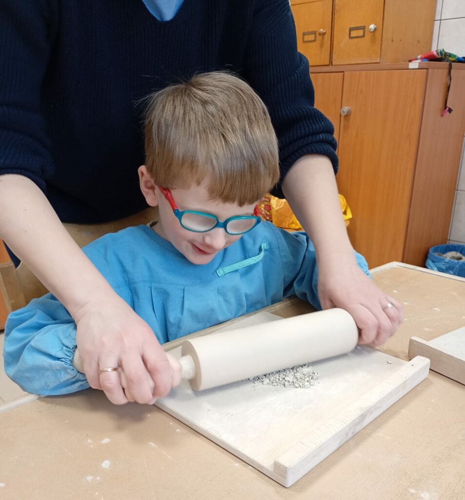 Uśmiechnięty chłopiec w fartuszku siedzi przy stole w pracowni ceramicznej. Przed nim na stole leży drewniana podkładka a na niej drobniutkie kawałki suchej gliny. Chłopiec z pomocą nauczycielki rozgniata kawałki gliny za pomocą wałka. 