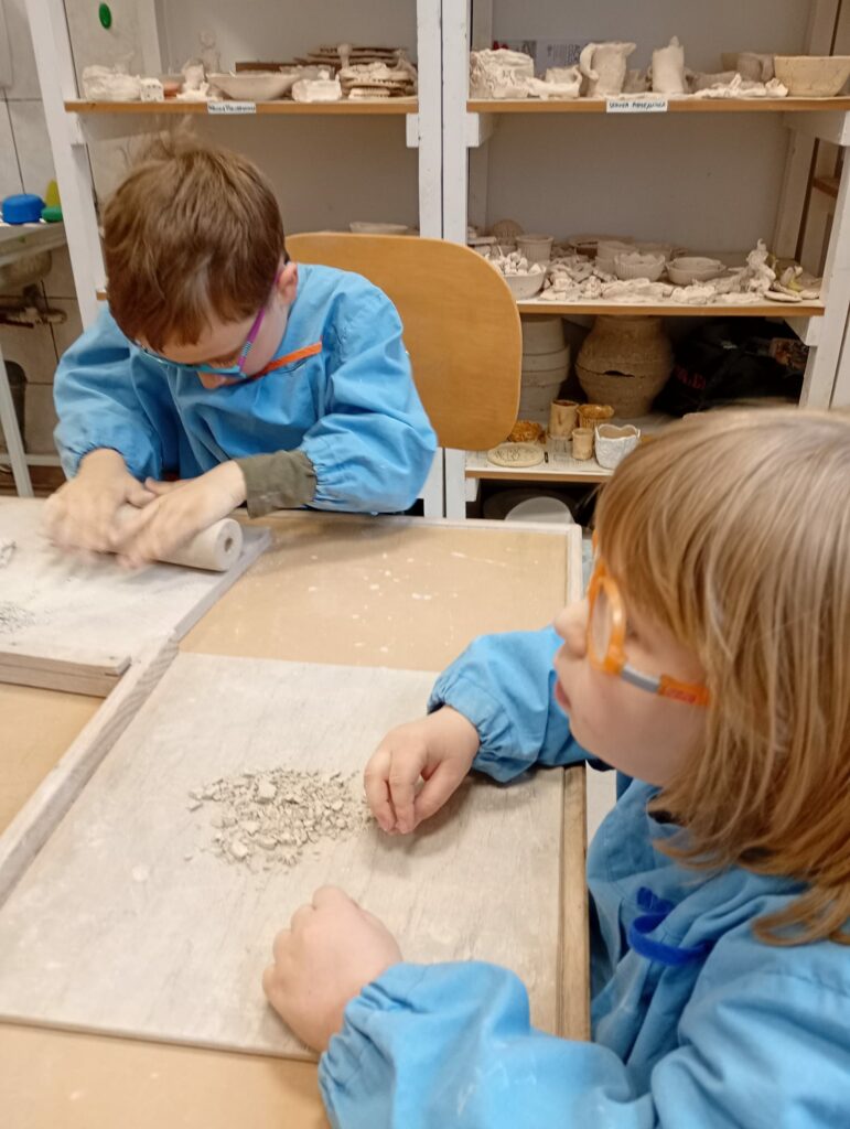 Chłopiec i dziewczynka w fartuszkach siedzą przy stole w pracowni ceramicznej. Przed każdym na stole leży drewniana podkładka, a na niej drobniutkie kawałki suchej gliny. Chłopiec rozgniata kawałki gliny za pomocą wałka. Z tyłu za chłopcem regały z półkami zastawionymi pracami z gliny.