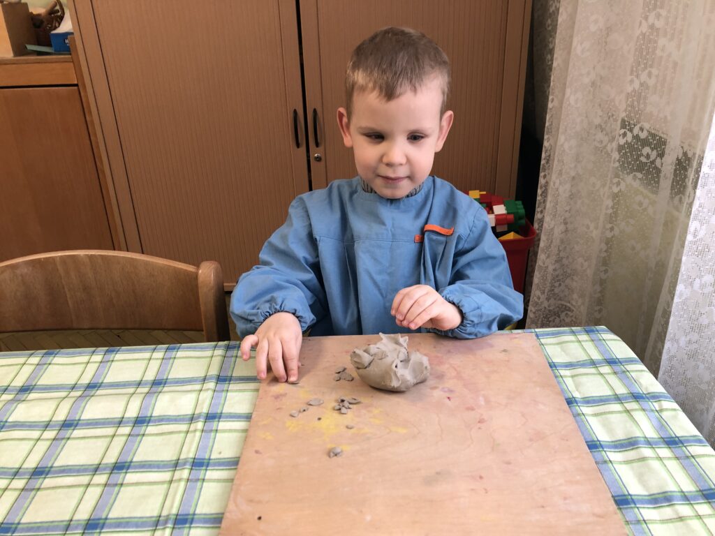 Uśmiechnięty chłopiec w fartuszku siedzi przy stoliku w sali przedszkolnej. Przed nim na stoliku leży drewniana podkładka a na niej kawałki gliny. Chłopiec palcami prawej dłoni przykleja malutki kawałek gliny do deseczki.