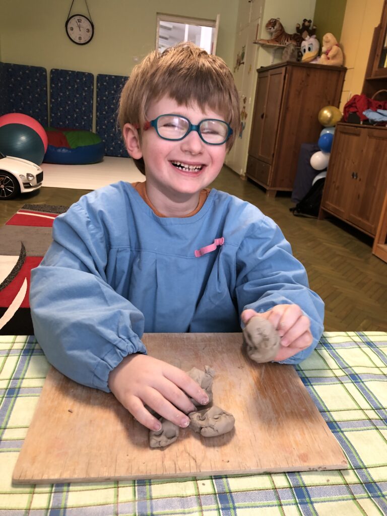 Roześmiany chłopiec w fartuszku siedzi przy stoliku w sali przedszkolnej. Przed nim na stoliku leży drewniana podkładka a na niej kawałki gliny. Chłopiec w lewej dłoni trzyma jeden kawałek gliny.