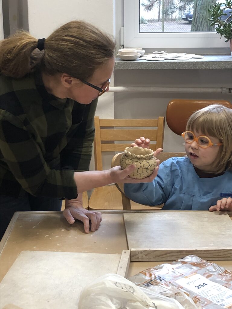 Dziewczynka w fartuszku siedzi przy stole w pracowni ceramicznej. Przed nią na stole leży drewniana podkładka. Dziewczynka patrzy i dotyka ceramicznego naczynia stojącego na dłoni nauczycielki stojącej obok dziewczynki. 