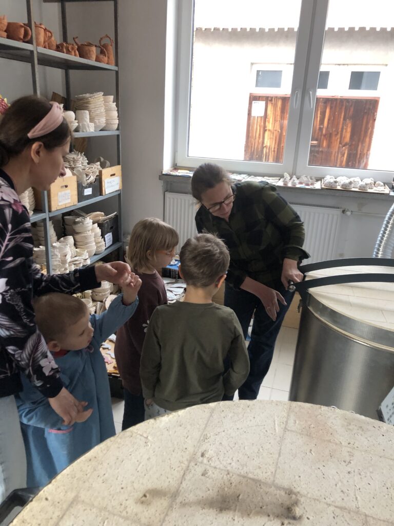 Trójka dzieci z nauczycielką stoi przy wielkim piecu w kształcie walca. Przodem do nich stoi druga nauczycielka, która trzyma rękę na uchwycie od przykrywy pieca. 