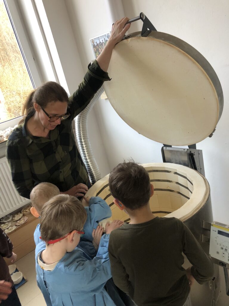 Trójka dzieci z nauczycielką stoi przy wielkim piecu w kształcie walca. Piec ma otwartą pokrywę. Nauczycielka trzyma rękę na uchwycie od pokrywy pieca. 