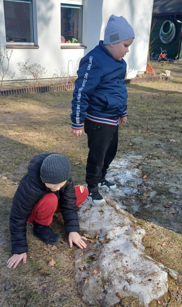 Jeden chłopiec stoi na małej górce śniegu. Drugi chłopiec przykuca i dotyka ręką resztek śniegu. 
