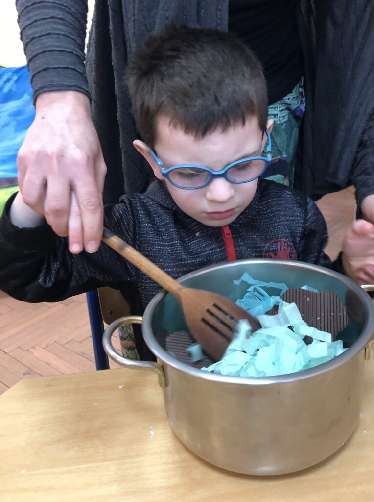 Chłopiec miesza drewnianą łyżką w metalowym garnku wypełnionym symbolami pogody.