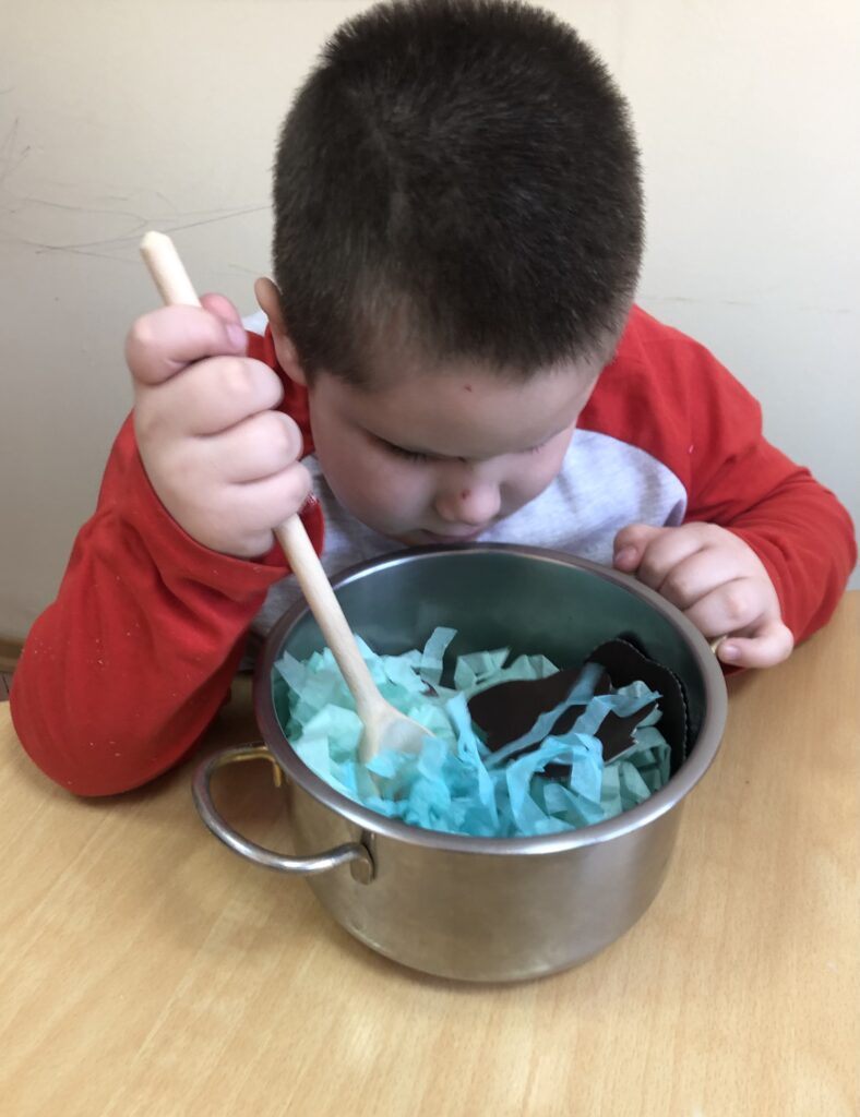 Chłopiec miesza drewnianą łyżką w metalowym garnku wypełnionym symbolami pogody.