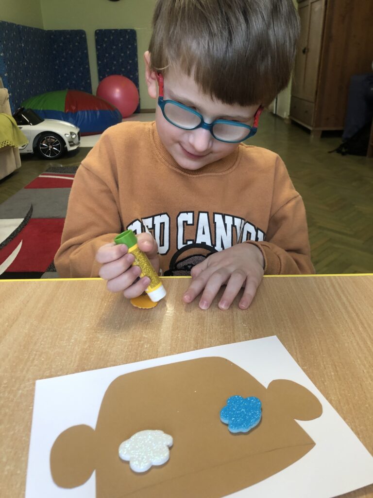 Chłopiec smaruje klejem żółte kółko. Obok leży kartka papieru z naklejonym modelem garnka, a na nim dwoma chmurkami.