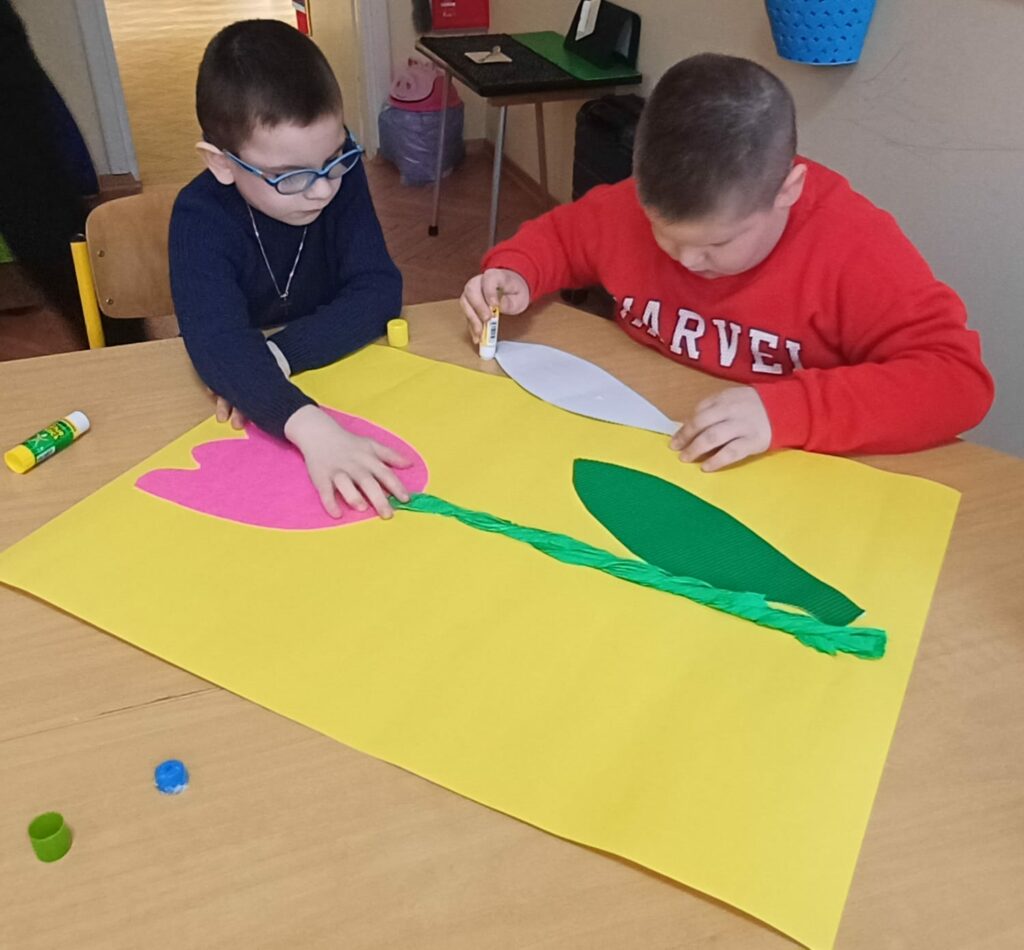 Jeden chłopiec smaruje klejem papierowy liść. Drugi chłopiec dotyka naklejonego papierowego kwiatka.