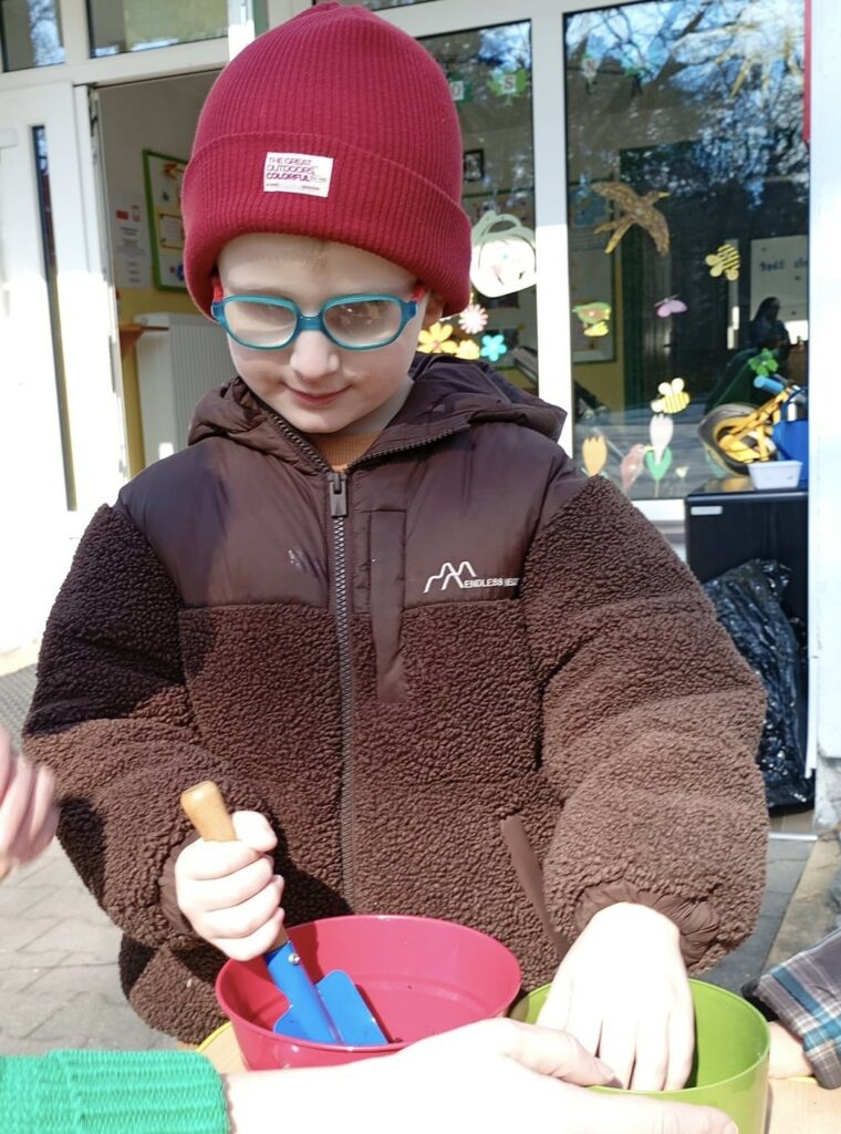 Chłopiec nasypuje łopatką ziemię z wiaderka do doniczki.