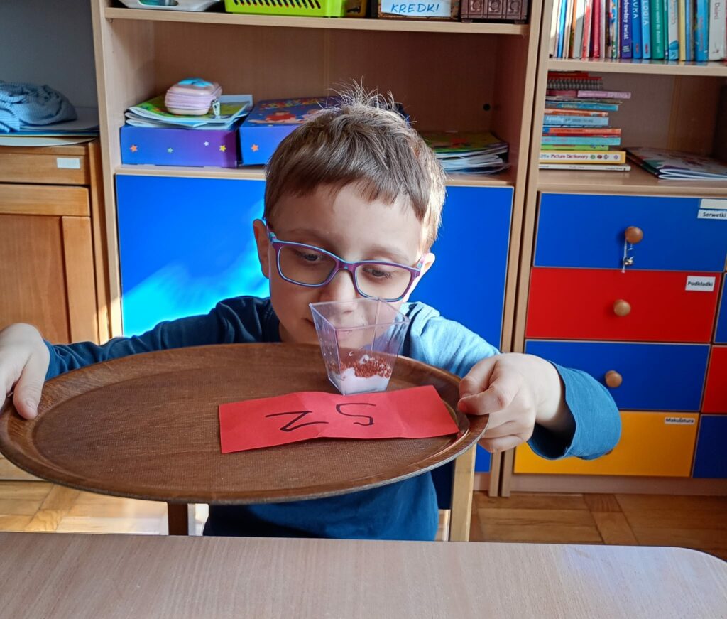 Chłopiec trzyma tacę. Na tacy stoi przezroczysty pojemnik z watą i nasionkami oraz kartka z napisem "sz".