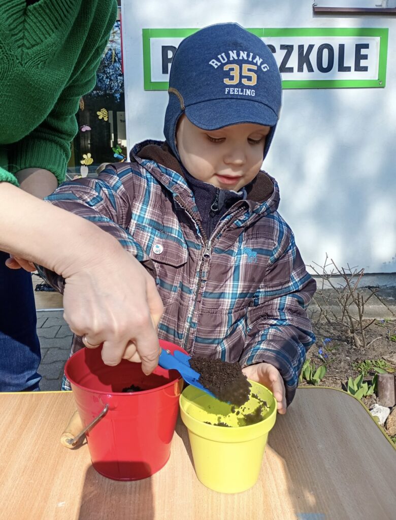 Chłopiec z pomocą nauczyciela nasypuje łopatką ziemię z wiaderka do doniczki.