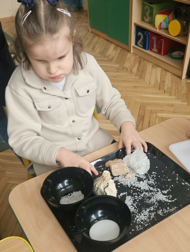 Dziewczynka ogląda białe i różowe bryłki soli leżące na czarnej tacy.