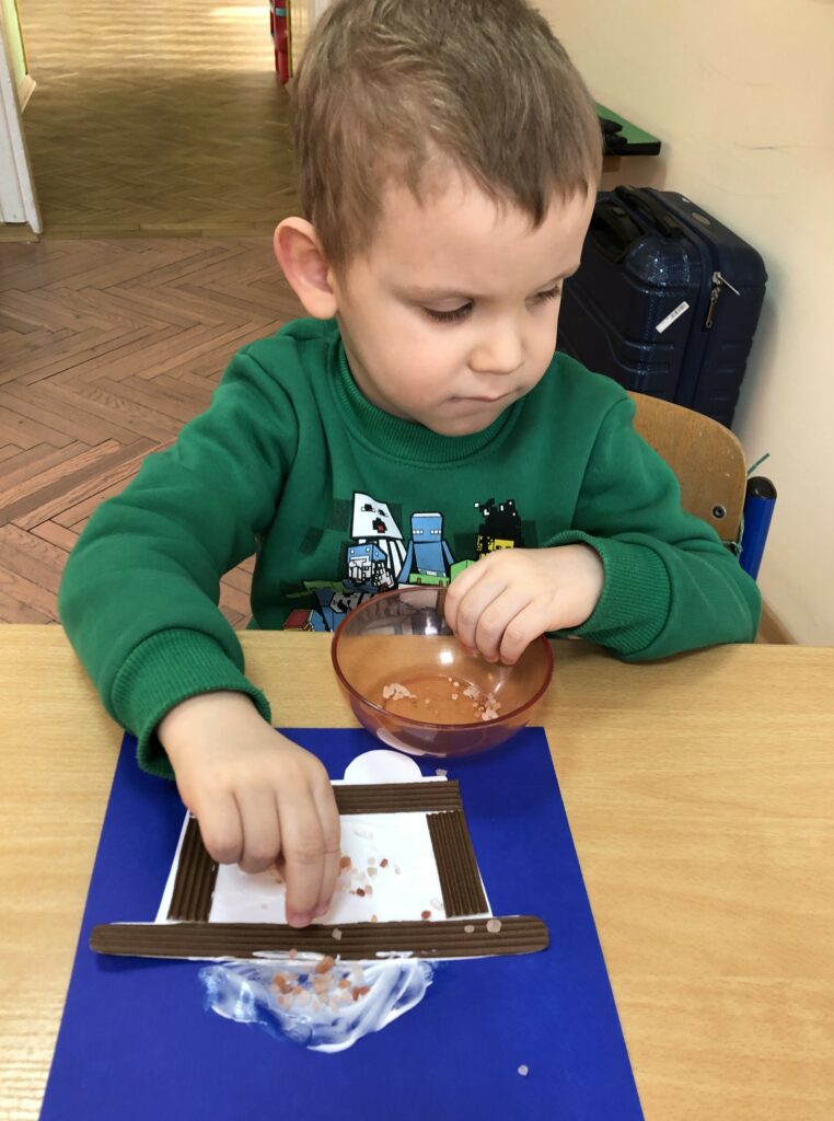 Chłopiec sypie małe kryształki soli na wyklejony na granatowej kartce wagonik z papieru.