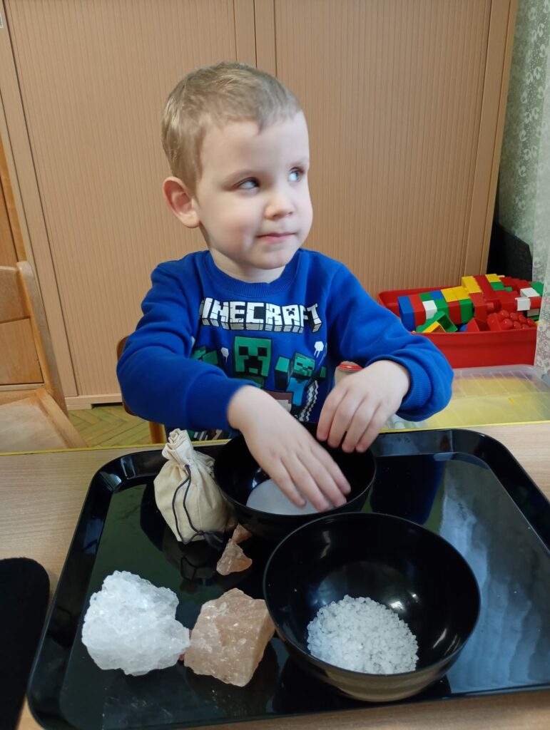 Na czarnej tacy znajdują się różnej wielkości bryłki soli, lniany woreczek z solą i 2 czarne miseczki z solą różnej granulacja. Chłopiec ogląda sól w jednej z miseczek. 