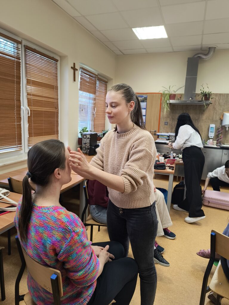 W jasnej sali warsztatowej uczennica w beżowym swetrze z ogromną uwagą i delikatnością wykonuje makijaż koleżance w kolorowym swetrze, ćwicząc w praktyce techniki manualne i precyzyjne nakładanie produktów.