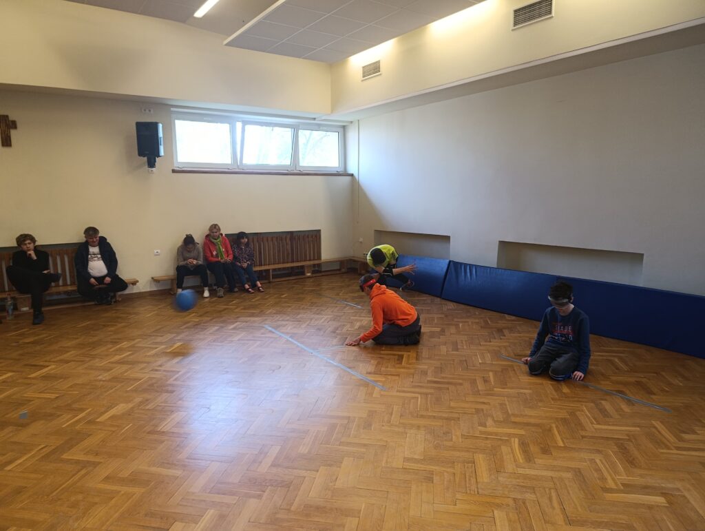 Uczniowie Szkoły Podstawowej w Laskach, z zasłoniętymi oczami i w pełnym skupieniu, rywalizują na parkiecie podczas emocjonującego turnieju w goalballa, śledząc słuchem ruch toczącej się piłki.