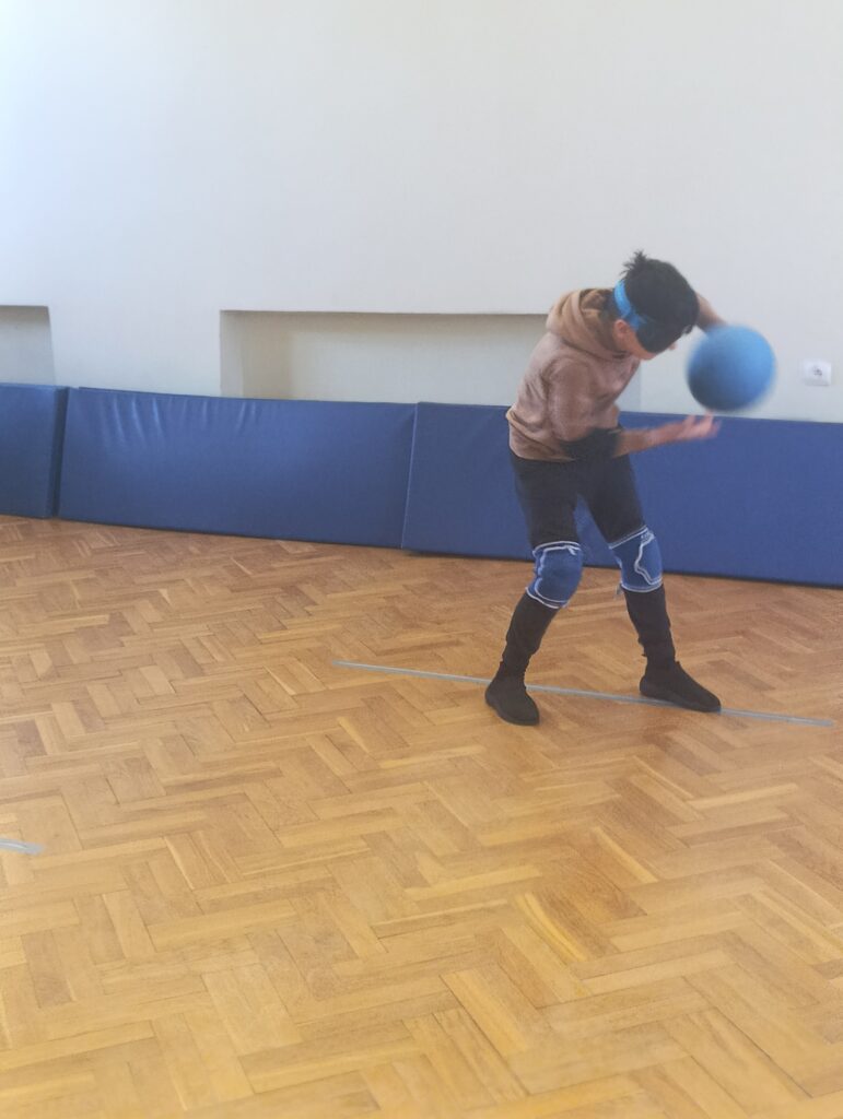 Zawodnik w goglach i ochraniaczach na kolana dynamicznie wyprowadza rzut piłką dźwiękową, celując w stronę bramki przeciwnika podczas szkolnego turnieju goalballa.