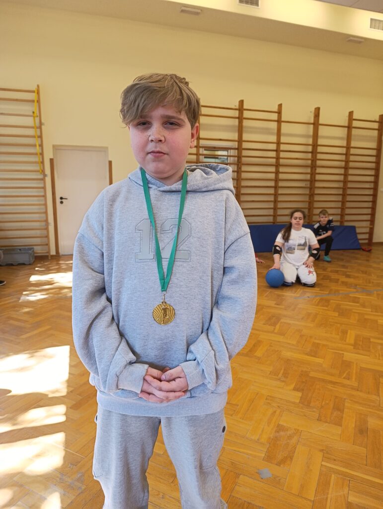 Młody zawodnik z dumą prezentuje na piersi złoty medal zdobyty podczas szkolnego turnieju goalballa, będący wyrazem uznania dla jego zaangażowania i sportowej postawy.