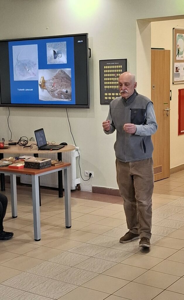 Paleontolog Michał Ginter, na tle prezentacji multimedialnej o budowie płaszczek, z zaangażowaniem omawia morfologię ryb chrzęstnoszkieletowych podczas wiosennego spotkania przyrodniczego w Zespole Szkół Ponadpodstawowych.