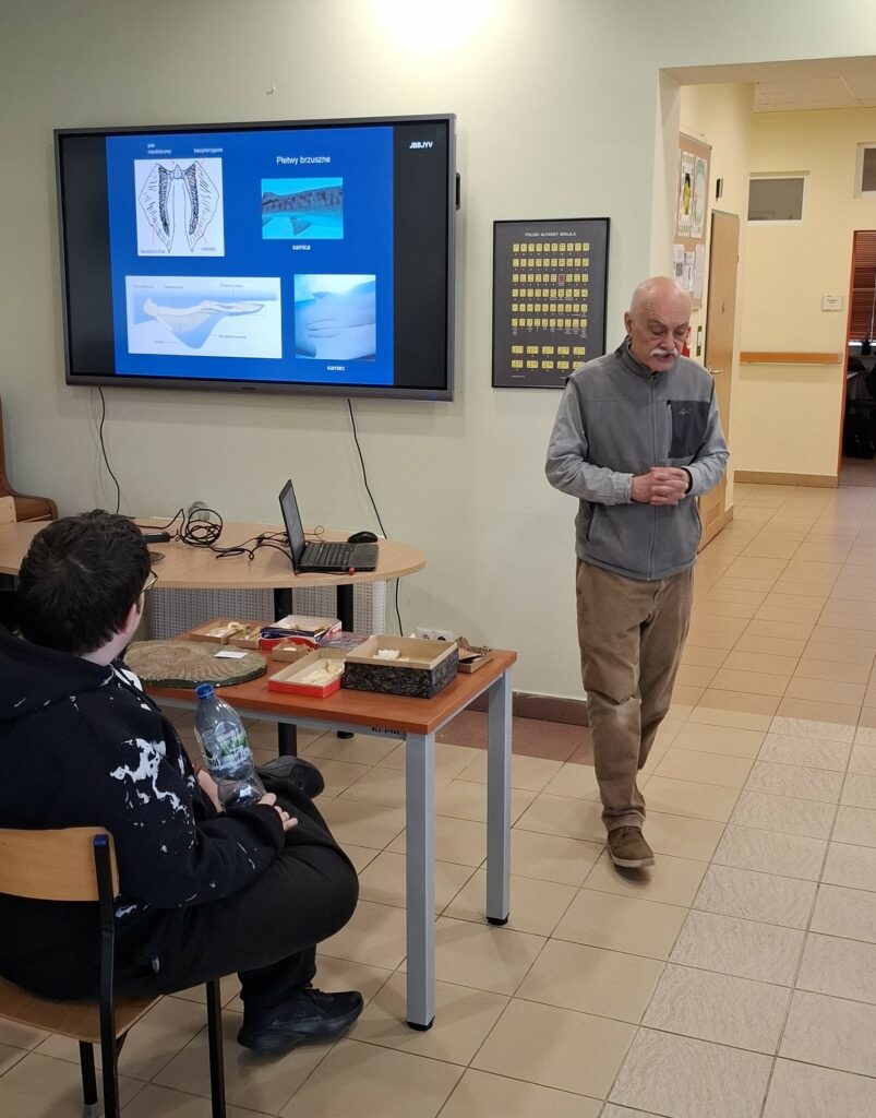 Podczas wiosennego spotkania przyrodniczego dr Michał Ginter prezentuje młodzieży anatomię płetw brzusznych rekinów i płaszczek, wyjaśniając różnice w ich budowie na przykładzie profesjonalnych schematów i zgromadzonych na stole eksponatów.