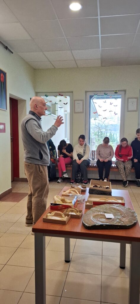 Pan Michał Ginter, paleontolog z Uniwersytetu Warszawskiego, z pasją opowiada zgromadzonej młodzieży o ewolucji morskich drapieżników, ilustrując swój wykład unikatowymi eksponatami szczęk i skamieniałości rozłożonymi na stole.