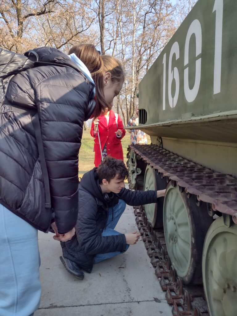 
Treść wiadomości Gemini
Uczniowie podczas plenerowej lekcji historii na terenie Cytadeli Warszawskiej szczegółowo badają układ jezdny zabytkowego pojazdu wojskowego. Chłopak przykuca przy gąsienicach i kołach nośnych, poznając ich konstrukcję poprzez dotyk, co pozwala mu na zrozumienie technicznych aspektów dawnego sprzętu militarnego