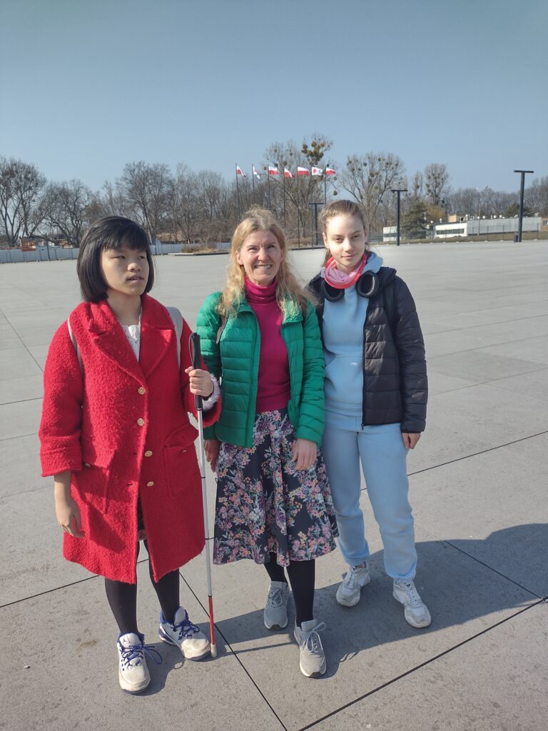 Trzy osoby stoją obok siebie na rozległym, nowoczesnym placu przed budynkiem Muzeum Historii Polski. Po lewej stronie znajduje się dziewczyna w czerwonym płaszczu trzymająca białą laskę, w środku kobieta w zielonej kurtce, a po prawej dziewczyna w niebieskim dresie i czarnej pikowanej kurtce z założonymi na szyję słuchawkami.