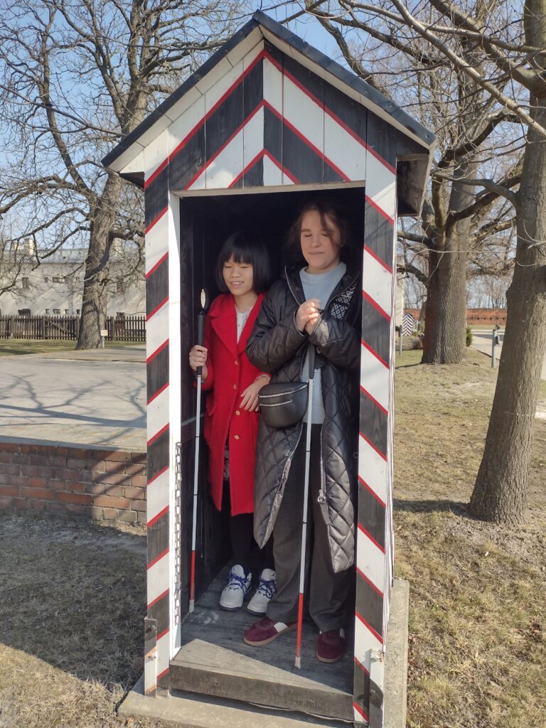 Dwie dziewczyny, obie trzymające białe laski, stoją wewnątrz drewnianej, historycznej budki wartowniczej na terenie Cytadeli Warszawskiej. Budka jest pomalowana w charakterystyczne, ukośne biało-czarno-czerwone pasy i ma spiczasty daszek.