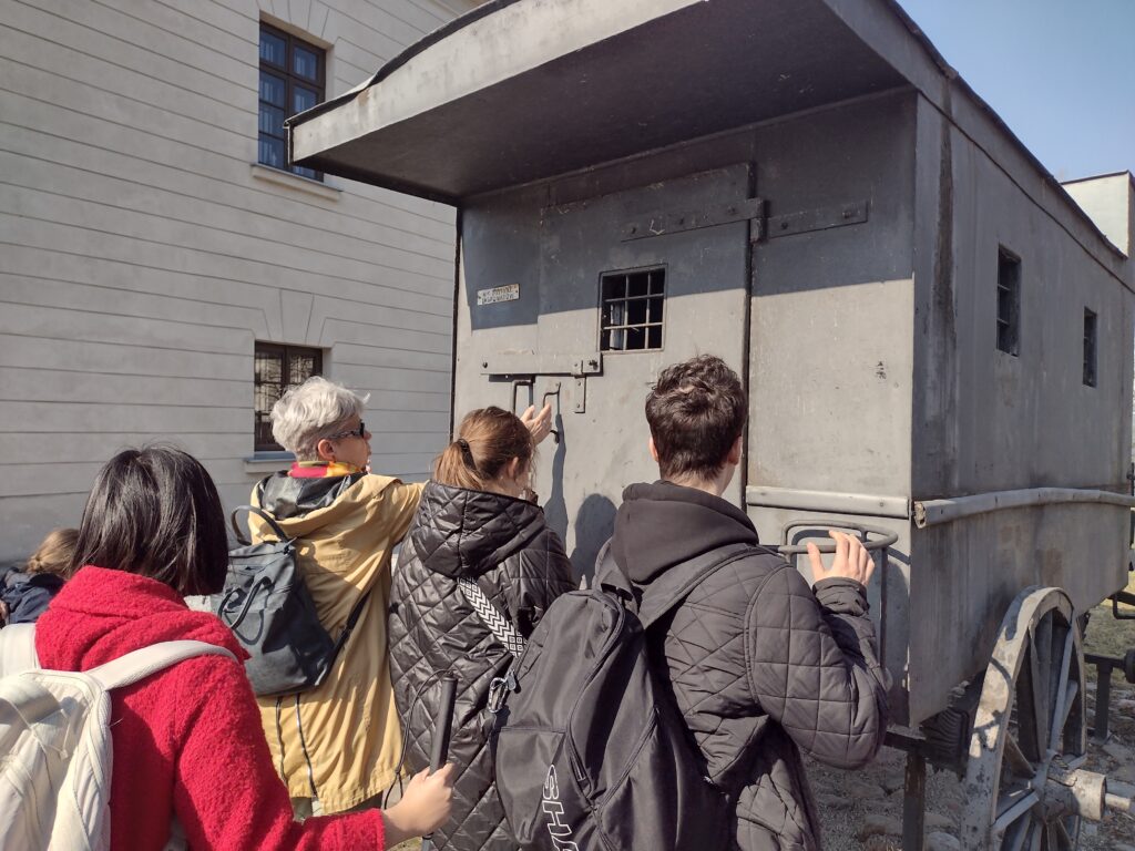 Młodzież podczas zwiedzania Cytadeli Warszawskiej ogląda z bliska historyczną więźniarkę konną, co pozwala im na bezpośredni kontakt z autentycznymi świadectwami przeszłości prezentowanymi na terenie muzeum.