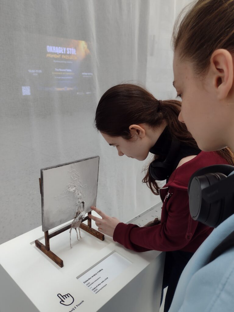 Uczennice w skupieniu poznają tyflografikę do pracy „Dziecko wojny z lalką” autorstwa Gero Hellmutha, która dzięki wypukłej formie pozwala na głęboki, dotykowy odbiór trudnej tematyki wystawy prezentowanej w Muzeum Historii Polski.