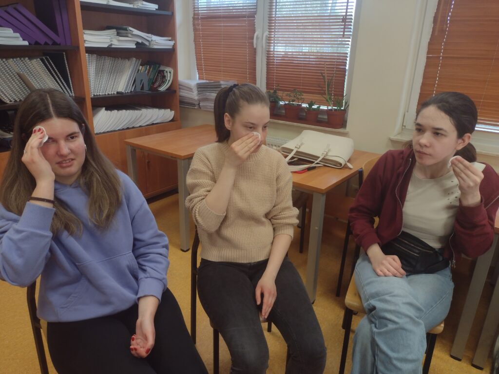 Trzy uczennice siedzą obok siebie i uważnie oczyszczają twarze płatkami kosmetycznymi, przygotowując cerę do dalszych etapów warsztatów makijażu pod okiem specjalistek.