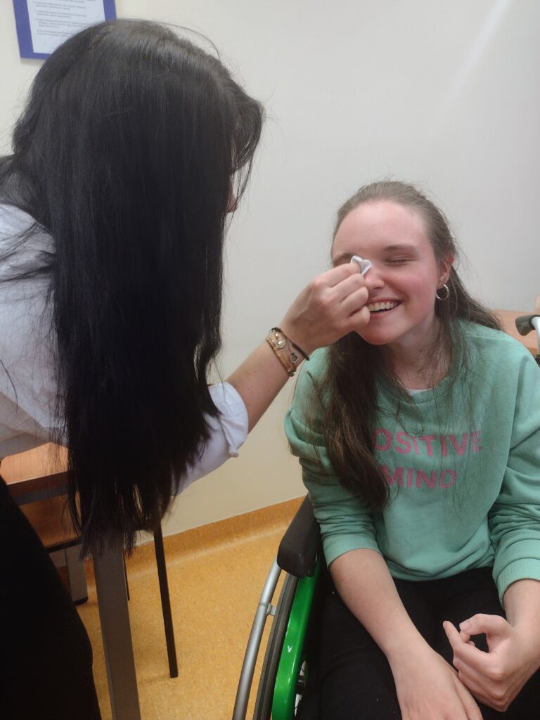 Makijażystka o długich, ciemnych włosach z delikatnością oczyszcza twarz płatkiem kosmetycznym uśmiechniętej uczennicy na wózku, przygotowując jej cerę do profesjonalnego makijażu w atmosferze pełnej radości.