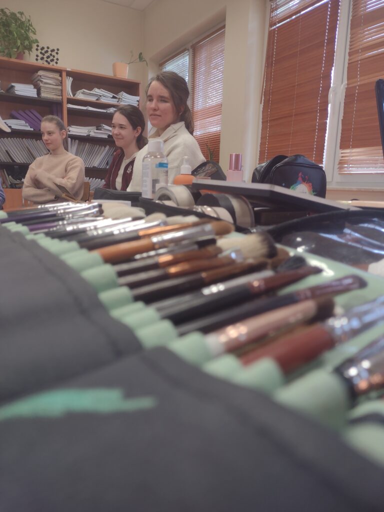Na pierwszym planie widać profesjonalny zestaw różnorodnych pędzli do makijażu, a w tle trzy uczennice, które z zaciekawieniem i uśmiechem obserwują pokaz, czekając na możliwość przetestowania tych narzędzi w praktyce.