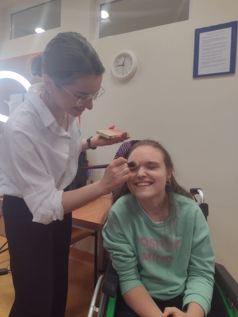 Makijażystka w okularach z uśmiechem pochyla się nad roześmianą dziewczyną na wózku inwalidzkim, nakładając jej pędzlem podkład na czoło i wspólnie ciesząc się pozytywną energią panującą podczas warsztatów.